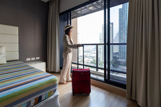 Asian woman moving the luxury hotel with luggage on her vacation while looking at the view from window with high rise building in the urban downtown area for business trip and condominium living usage