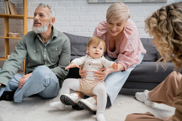 happy middle aged woman playing with little granddaughter near family in living room.