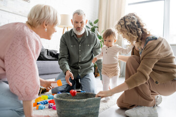 blonde woman with toddler daughter and mature parents near wicker basket and toys on floor in living room.