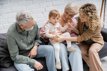 smiling woman holding hands of toddler daughter while spending time with parents on couch at home.