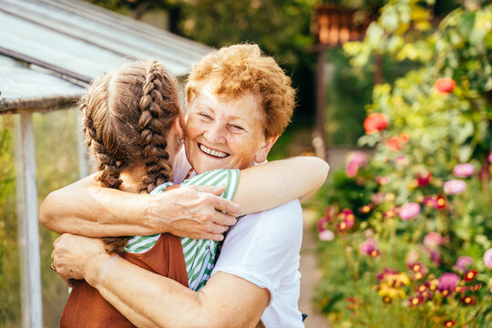 Rear view of young teenager girl with cute hairstyle two braids hugging her happy loving grandmother at summer countryhouse outdoor.