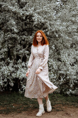 A young beautiful red-haired woman stands near a branching spring apple tree. A girl in a white dress smiles between blossoming apple trees. Spring season concept.