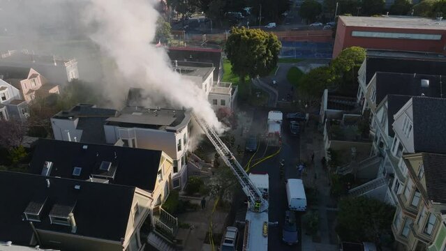 Aerial view of scene of house fire on street in residential area of city. Firefighters extinguish blaze from fire truck ladder.