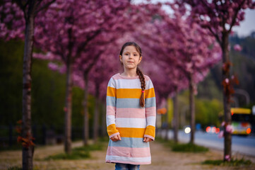 Fototapeta premium Little preschooler girl standing in striped dress before of blooming pink sakura alley