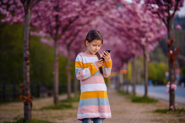 Little preschooler girl standing in striped dress before of blooming pink sakura alley