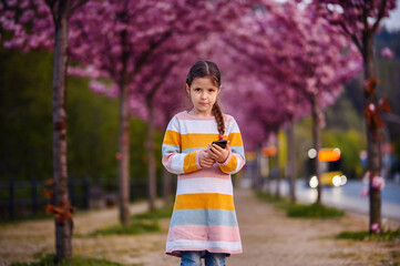 Little preschooler girl standing in striped dress before of blooming pink sakura alley