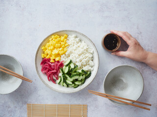Hand placing bowl of soy sauce on the table as a dressing for homemade poke bowl of rice, mango, cucumber and pickled red onion.