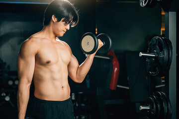 Fototapeta na wymiar An Asian man, shirtless and sweaty, holds heavy dumbbells, working his shoulder and bicep muscles in a dark gym for his fitness training. Fitness GYM Healthy sports lifestyle concept