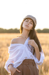 Fototapeta na wymiar Sensual portrait of a girl in a man's shirt and cap in a field at sunset with an ear of wheat
