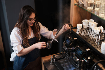 Barista standing next to a coffee machine in a coffee shop and heating up milk.