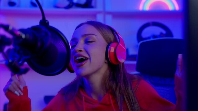 Portrait of young singer woman singing in headphones sitting at desk in music record home studio with neon light, Live or streaming cover singing or recording voice advertising concept