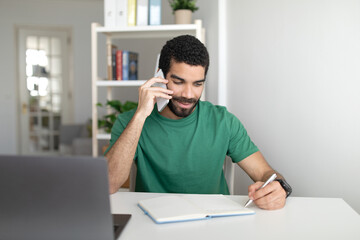 Smiling young middle eastern guy manager at table with laptop working remotely, call by smartphone