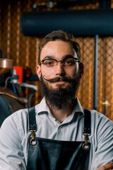 smiling handsome young man in apron and glasses stands in coffee production near machine for roasting coffee beans