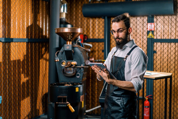 Fototapeta na wymiar serious guy in apron standing in coffee production near machine for roasting coffee beans with tablet in hands