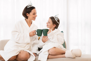 Little Girl And Her Mother Wearing Bathrobes And Crowns Relaxing At Home