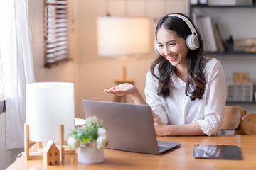Attractive happy young Asian businesswomen at home, sitting at the desk, using a laptop computer, tablet and headphones having a video chat.