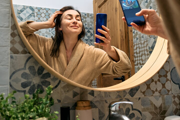 Middle-aged woman in bathrobe looking at mirror in bathroom, touching her face, smiling and using...