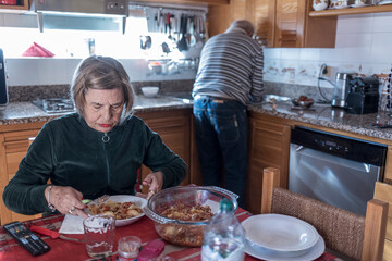 signora anziana mangia seduta al tavolo della cucina, mentre il marito si adopererà a portare il cibo in tavola