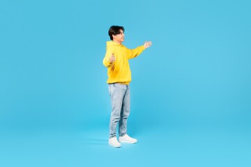 Fototapeta na wymiar Positive Korean Teenager Guy Spreading Hands Looking Aside, Blue Background