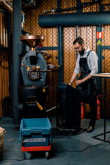 a handsome young bearded man in an apron enters data into a machine for roasting coffee beans at a factory