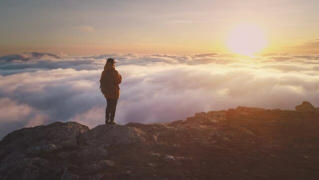 Tourist girl backpacker stands with backpack above the cloudy sky and looks at sunset