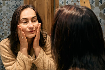 Middle-aged woman in bathrobe looking at mirror in bathroom and touching her face, applying...