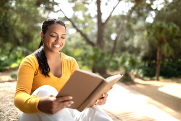 Smiling brazilian student lady reading book outdoors, preparing for lectures or exams while sitting in park, copy space