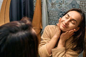 Middle-aged woman in bathrobe looking at mirror in bathroom and touching her face, applying hydrating anti-aging cosmetics on her skin and smiling.