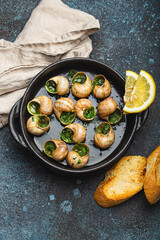 Escargots de Bourgogne Snails with Garlic Butter and Parsley in black cast iron pan with Lemon and Toasted Baguette Slices on rustic stone background top view, traditional French Delicacy .