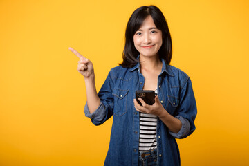 Portrait beautiful young asian woman happy smile dressed in denim jacket showing smartphone with pointing finger hand gesture to free space isolated on yellow studio background. app smartphone concept - obrazy, fototapety, plakaty