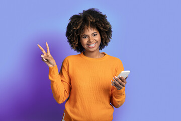 Happy young black woman using smartphone, showing peace gesture