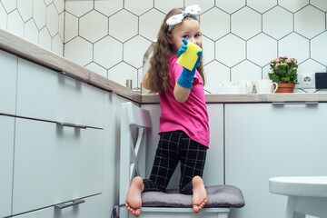 Portrait of wonderful little girl with long dark hair wearing pink T-shirt, black leggings, big blue gloves, sitting on stool near sink, holding yellow sponge in kitchen, looking back. Dishwashing.
