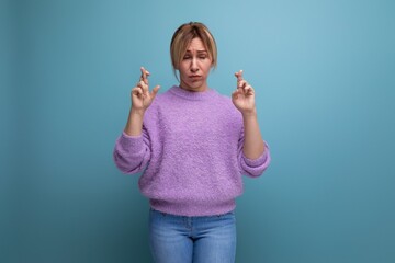 portrait of hopeful charming pleasant blond young woman in purple sweater crossing fingers on blue background with copy space