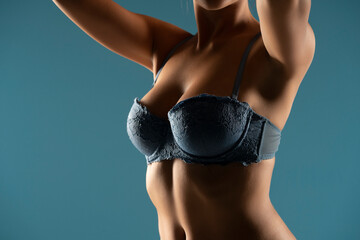Close up on woman torso in the shadow. Blue bra and natural breasts cleavage on a blue background - obrazy, fototapety, plakaty