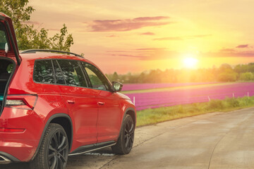 Modern red SUV car parked on rural road near blooming flower field against scenic warm evening sunset light sky landscape. Family countryside vacation auto journey trip at nature outdoors