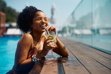 Happy black woman drinks cocktail at pool on summer vacation.