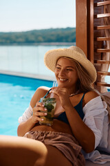Young woman enjoys in summer cocktail while relaxing on deck chair by pool.