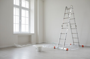 Modern home after repairs and renovations. New empty house interior with white freshly painted walls, step ladder, paint can, protective plastic on the floor, big window, and no people