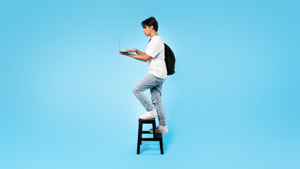 Fototapeta na wymiar Asian Student Guy Using Laptop Walking Up Stairs, Blue Background