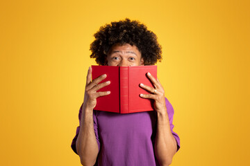 Cheerful excited adult african american curly man in purple t-shirt looks from behind book