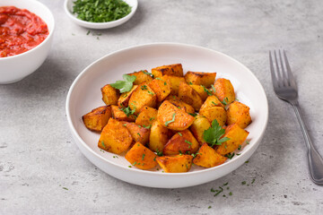 Traditional Spanish potato, patatas bravas with smoked paprika, spicy tomato sauce and parsley in a white bowl on a gray stone background, top view. Delicious homemade food
