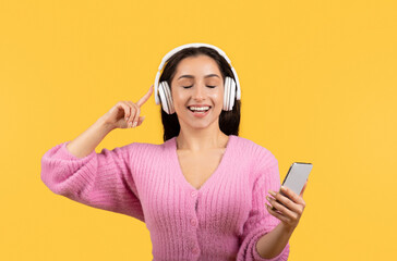 Cheerful millennial arab lady in casual and wireless headphones listen music, sings song with smartphone