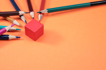 A red wooden cube on the orange background with colorful pencils at the side. Copy space for text and messages 