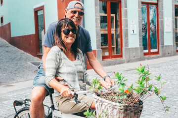 Smiling cheerful senior couple sitting outdoors on the same bicycle enjoying a sunny day in a street city. Healthy lifestyle and retirement concept