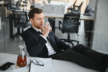 Fatigue young businessman sitting at workplace and holding whiskey bottle, drinking alcohol, bankruptcy concept.
