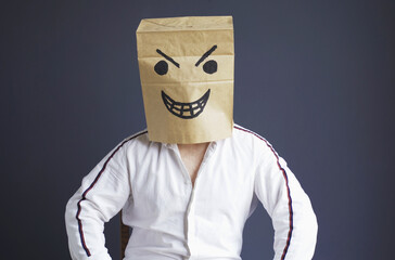 A man with a paper bag on his head, with a drawn angry smiley face, sits in a white chair. - obrazy, fototapety, plakaty