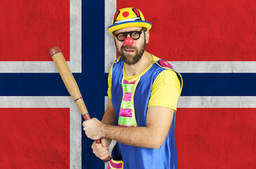 An insecure clown holds a bat in his hands against the background of the flag of Norway