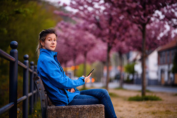 Young girl in blue jacket and jeans sitting on bench on street with sakura alley using smartphone