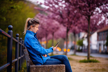 Young girl in blue jacket and jeans sitting on bench on street with sakura alley using smartphone