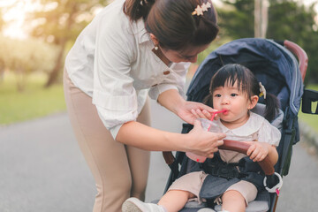 Obraz premium Asian mother walking with daughter in stroller walking in park, Mom feed water to child.